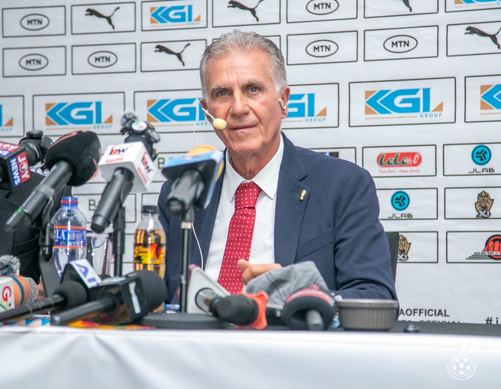 Male official in a dark suit and red tie speaks at a press conference, with multiple microphones in front and sponsor logos on a white backdrop behind him. Male official in a dark suit and red tie speaks at a press conference, with multiple microphones in front and sponsor logos on a white backdrop behind him.