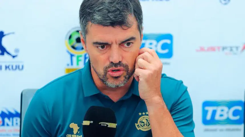 Man in a teal polo speaks into a microphone at a press conference, touching his ear with his hand. Man in a teal polo speaks into a microphone at a press conference, touching his ear with his hand.