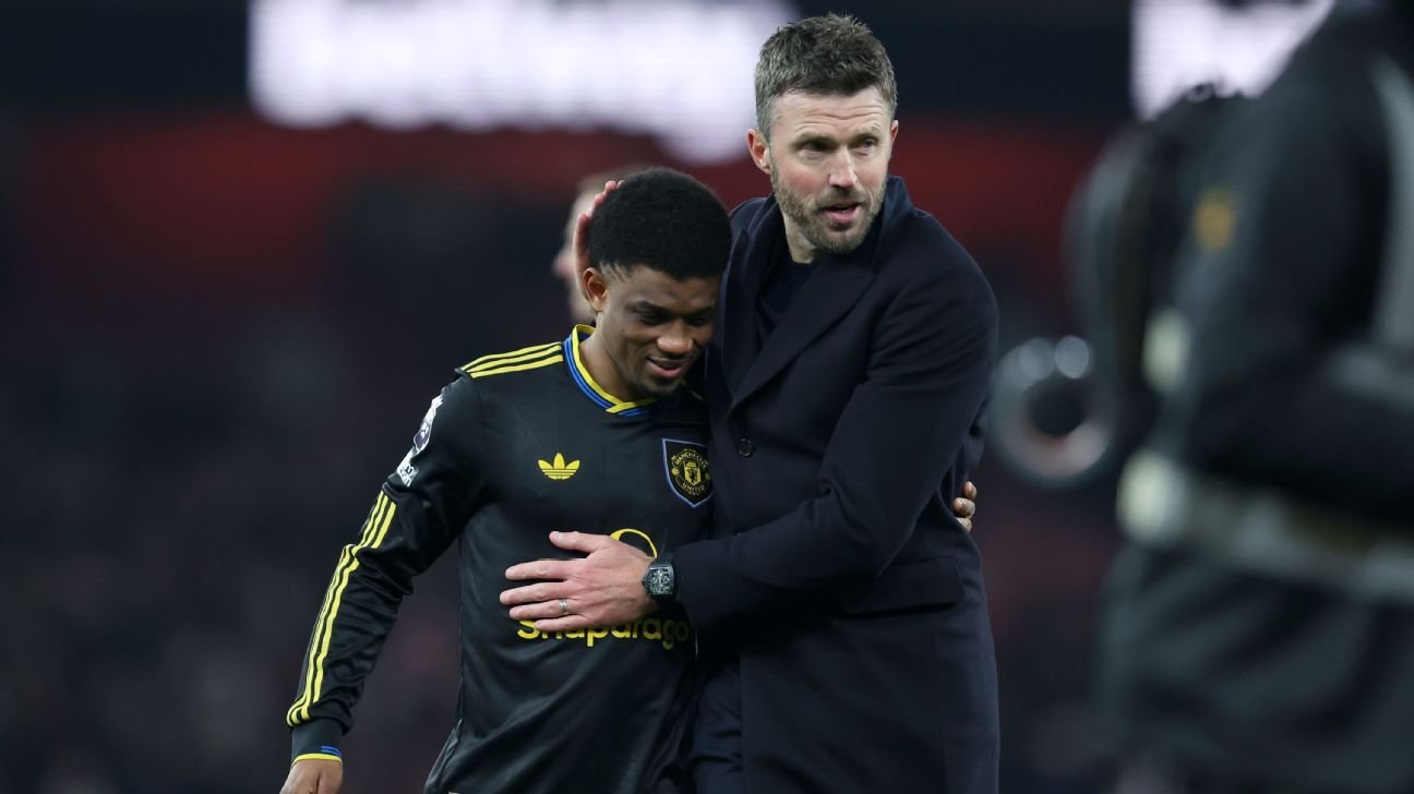 Two men share a hug on a soccer field; one in a black jersey with yellow trim, the other in a dark coat, both looking downcast.