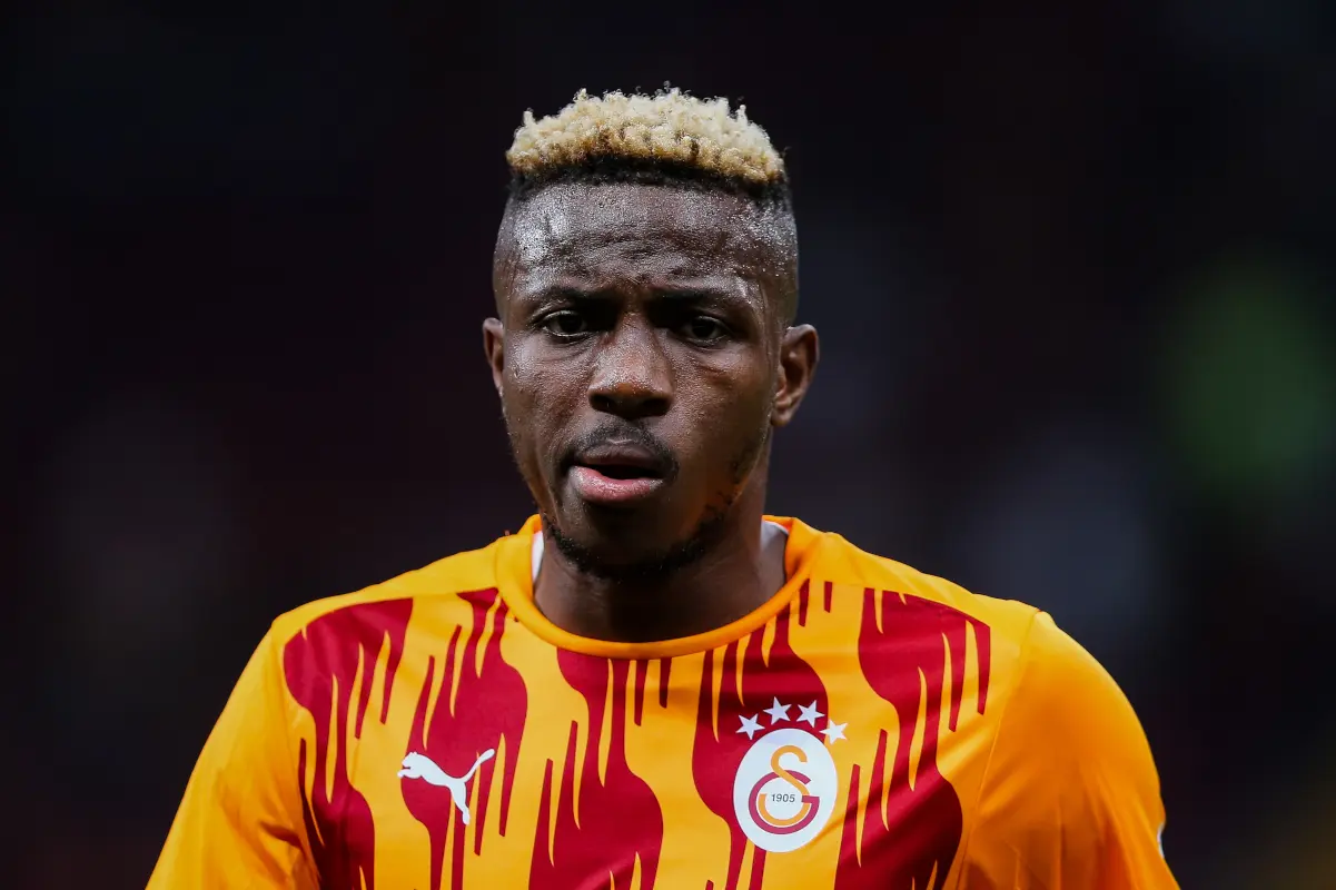 Galatasaray soccer player on the field, wearing a yellow-orange jersey with red flame patterns and the club crest, looking focused amid a dark stadium background.