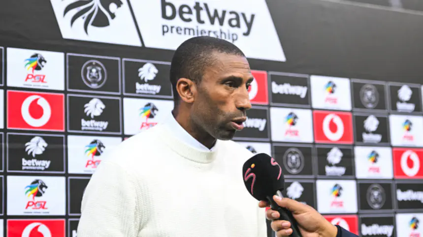 Man in a white sweater being interviewed, holding a microphone, with sponsor boards behind him.