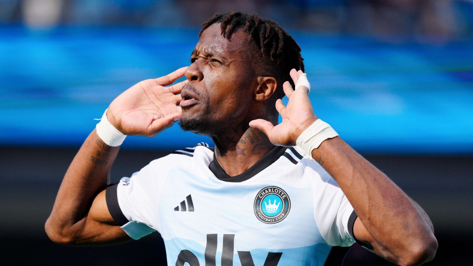 Soccer player in a Charlotte FC jersey cups his ears with hands during a match, celebrating loudly.