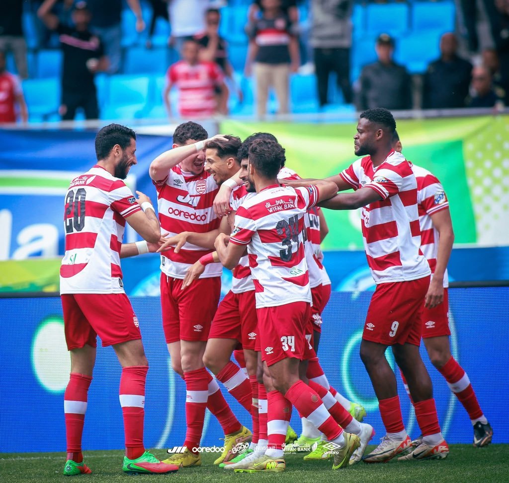 Soccer players in red-and-white striped uniforms celebrate in a huddle on the field, arms around each other, with blue stadium seats in the background