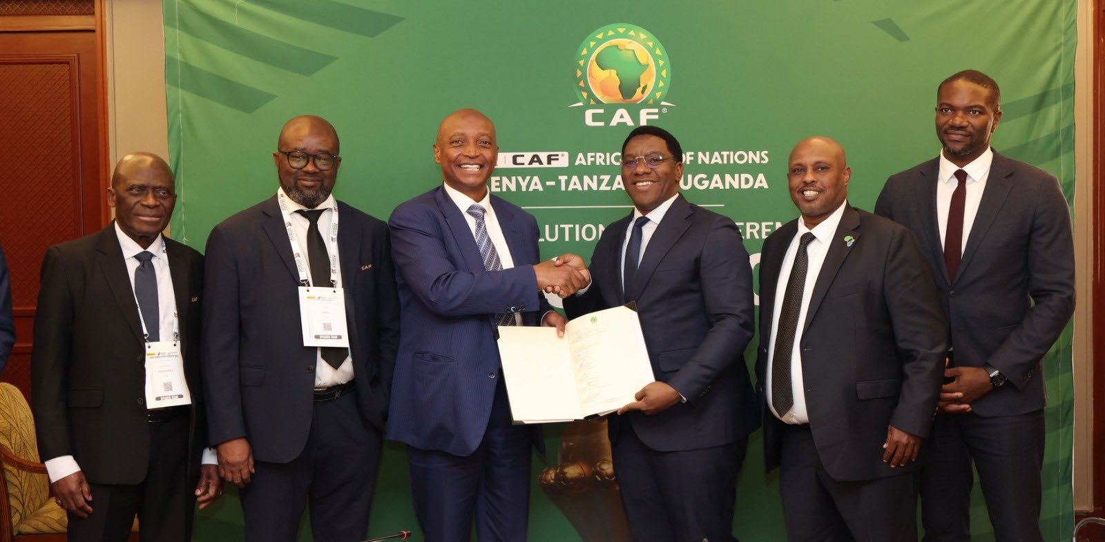 Six men in suits posing for a ceremony, two in the center shake hands and hold an open document against a green CAF banner backdrop. Six men in suits posing for a ceremony, two in the center shake hands and hold an open document against a green CAF banner backdrop.
