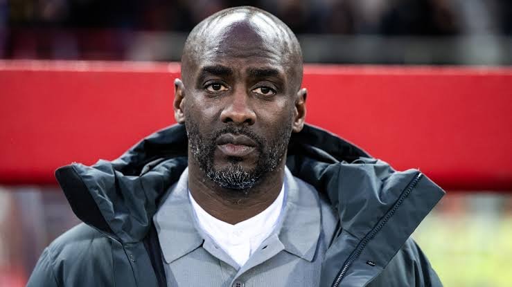 Portrait of a bald Black man with a gray beard, wearing a gray shirt and dark jacket, standing in front of a red barrier. Portrait of a bald Black man with a gray beard, wearing a gray shirt and dark jacket, standing in front of a red barrier.