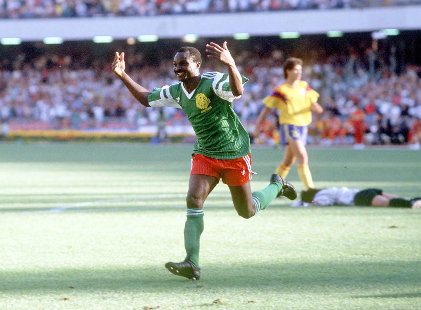 Soccer player in green jersey and red shorts celebrates with arms raised on a sunny field, crowd cheering in the stands behind him. Soccer player in green jersey and red shorts celebrates with arms raised on a sunny field, crowd cheering in the stands behind him.