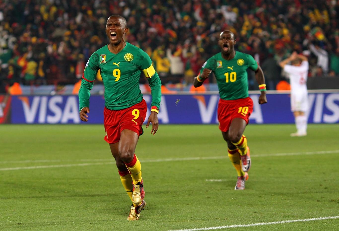 Two Cameroon players celebrate a goal on the pitch, wearing green jerseys with numbers 9 and 10 as the crowd cheers. Two Cameroon players celebrate a goal on the pitch, wearing green jerseys with numbers 9 and 10 as the crowd cheers.