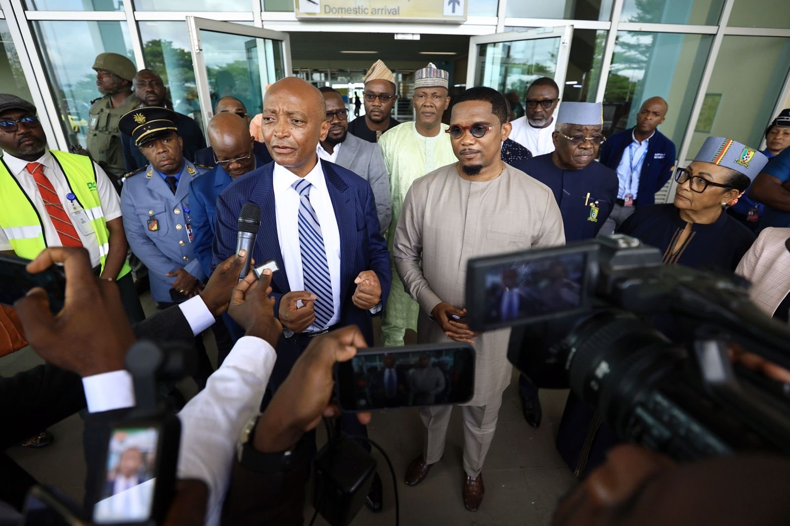 A man in a blue pinstripe suit speaks into a microphone while reporters, officials, and security surround him outside a terminal labeled 'Domestic arrival'.