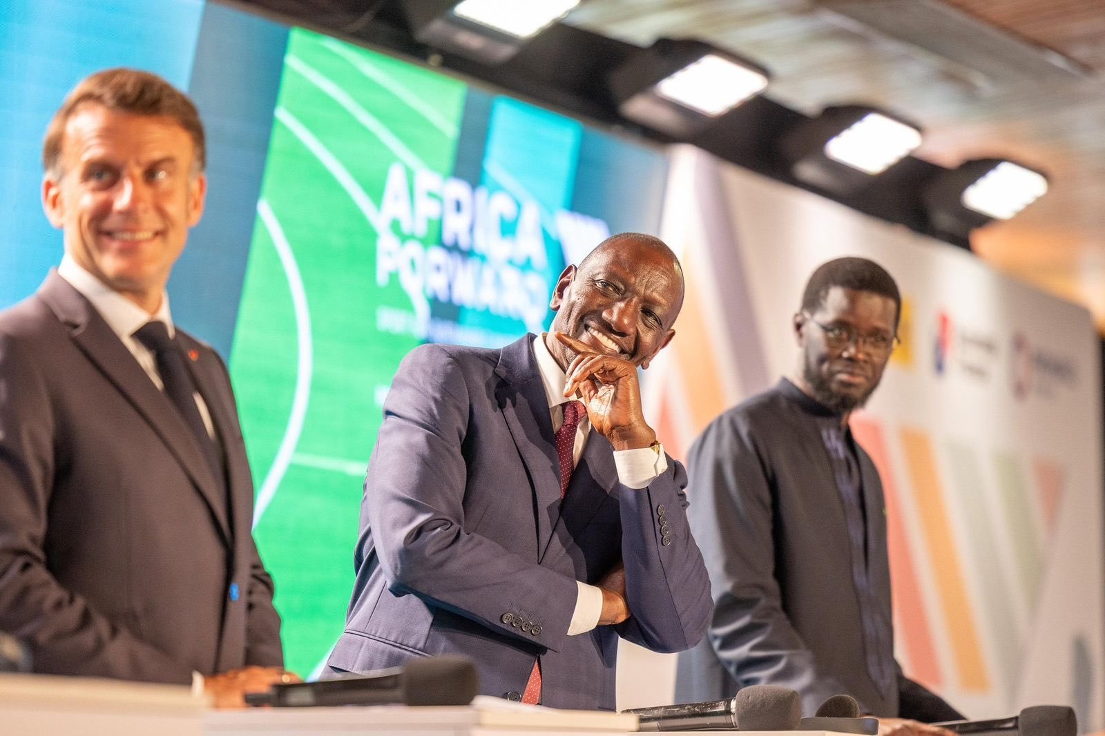Three men in suits stand at a conference panel; the center man smiles with his hand to his chin, as a green backdrop reads  Three men in suits stand at a conference panel; the center man smiles with his hand to his chin, as a green backdrop reads'Africa Forward' behind them.