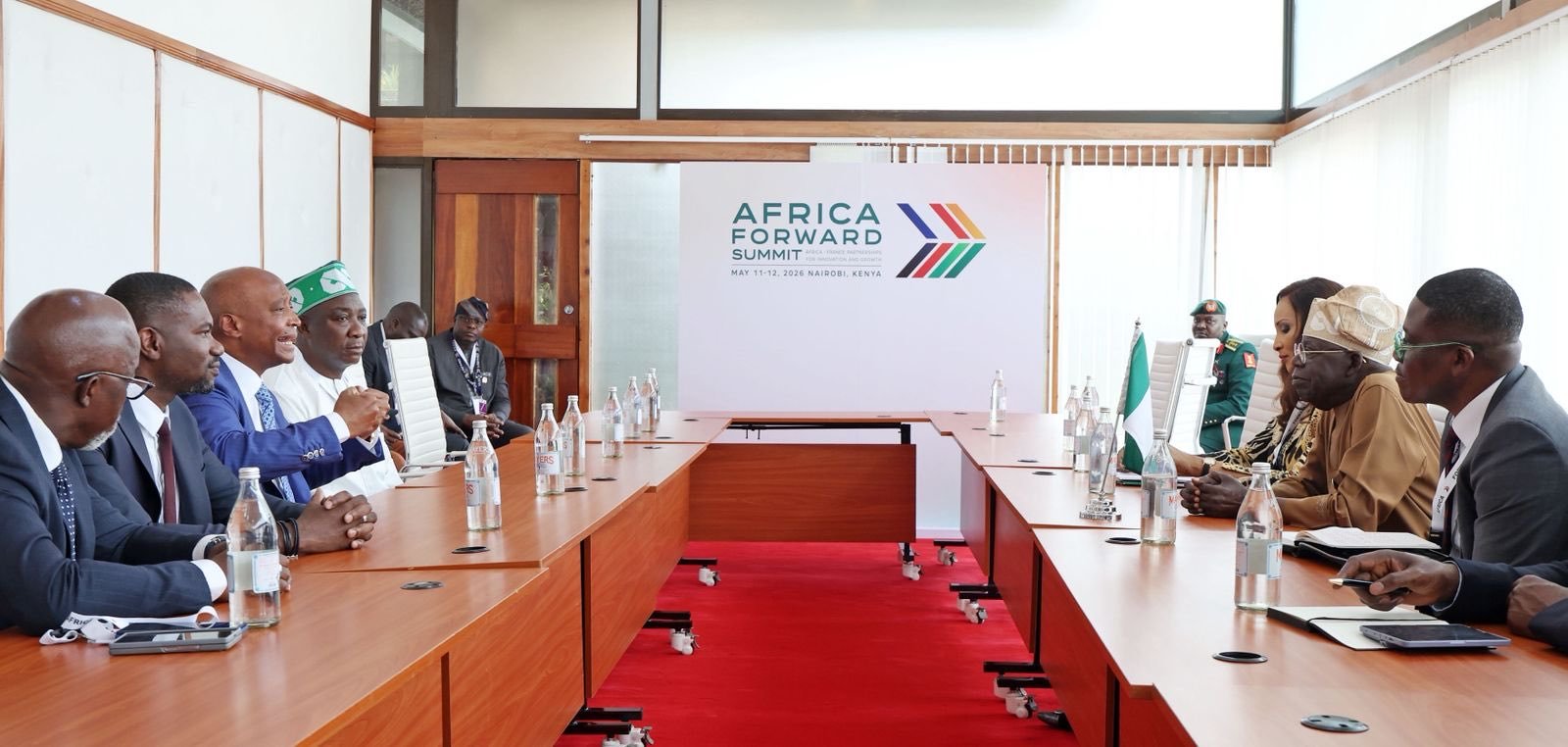 A formal meeting in a bright conference room with delegates seated along a U-shaped wooden table, facing a banner that reads 'Africa Forward Summit' with colorful arrows. Bottled water is on the table. Rules and flags are visible in the background.