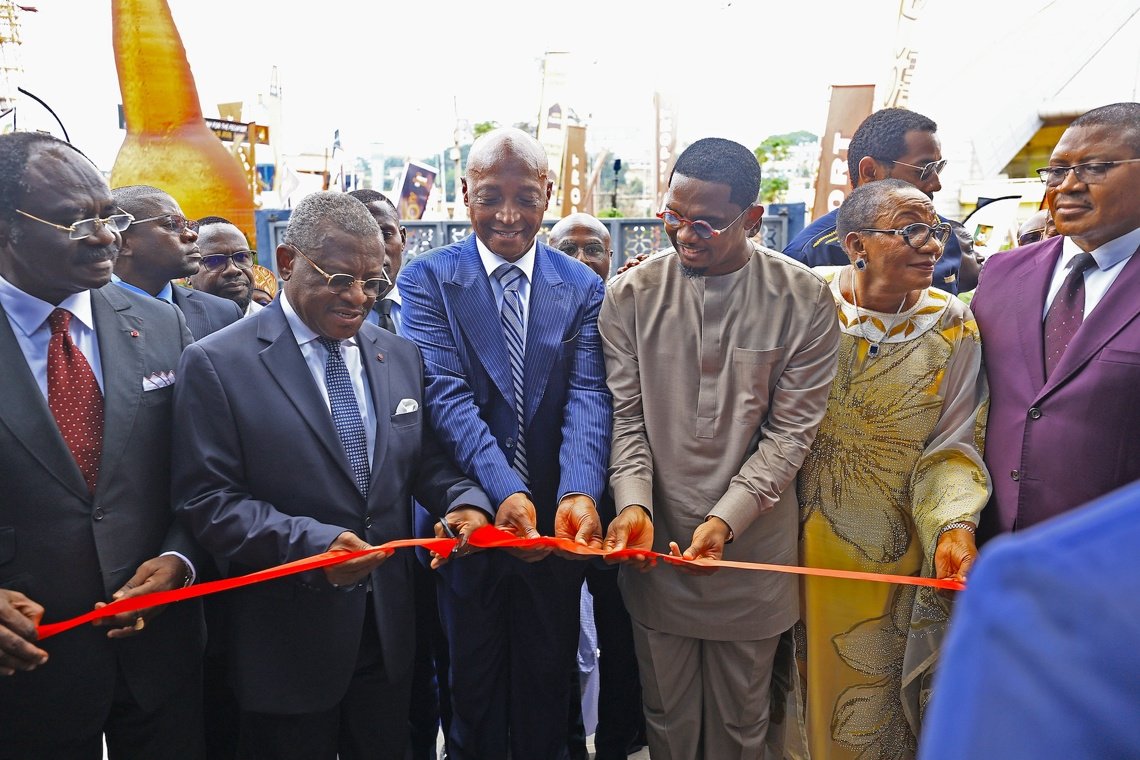 Group of officials cutting a red ribbon at a ceremony, with suited men and a woman in traditional attire watching smilingly behind them Group of officials cutting a red ribbon at a ceremony, with suited men and a woman in traditional attire watching smilingly behind them