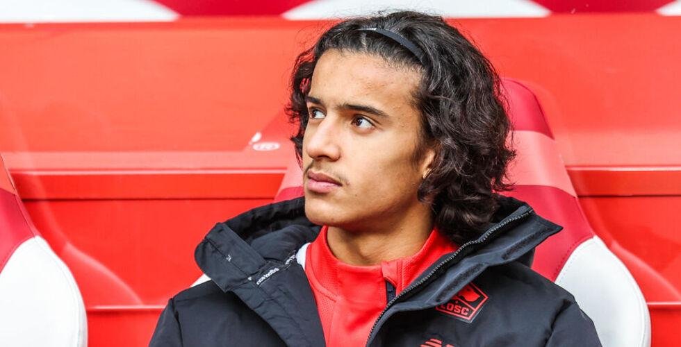 Young man with shoulder-length dark hair, wearing a black jacket over a red shirt, seated in red stadium seats and looking to the right. Young man with shoulder-length dark hair, wearing a black jacket over a red shirt, seated in red stadium seats and looking to the right.