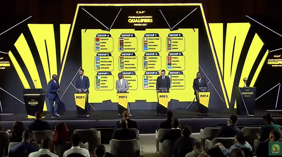 Stage during a CAF Africa Cup qualifiers draw ceremony; five presenters stand at podiums labeled POT 1–POT 4, facing an audience, with a large yellow display listing Groups A–L and country flags behind them.