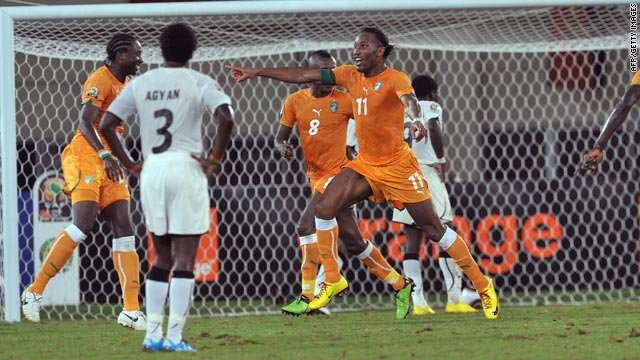 Soccer players in orange celebrate a goal at the goal net as teammates point and interact with players in white kits.