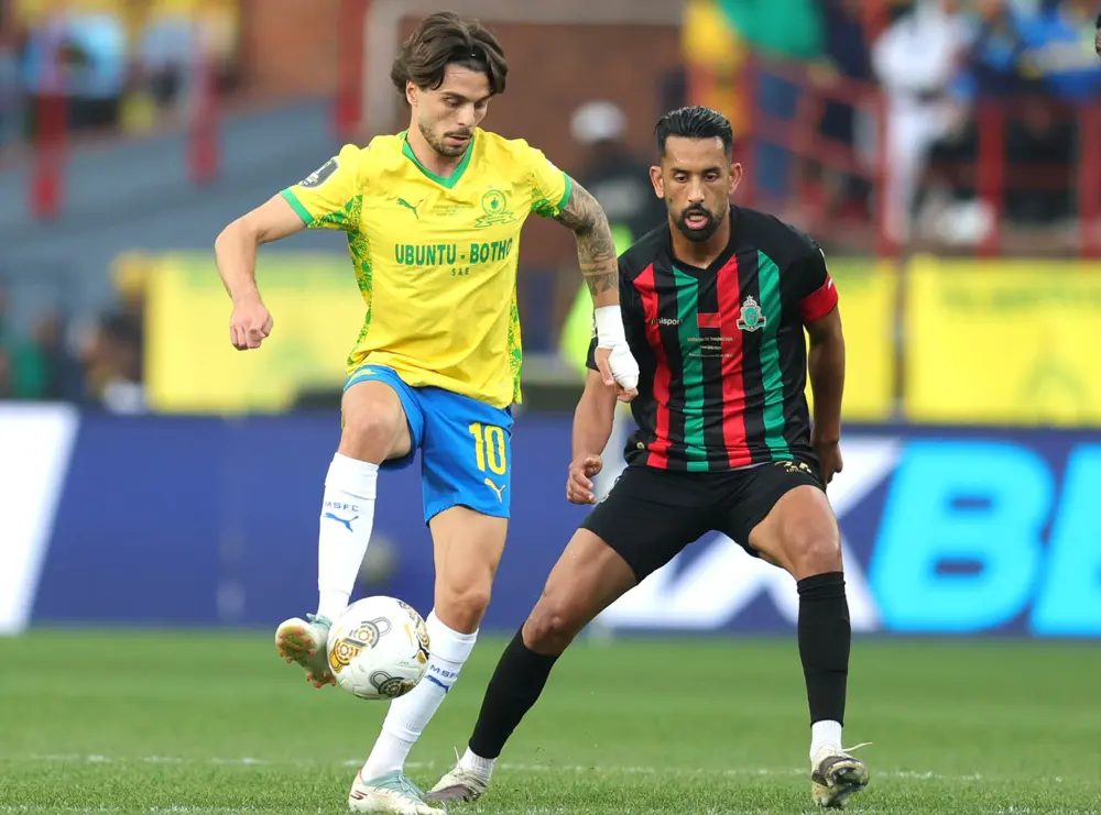 Two soccer players chase the ball during a match: one in a yellow jersey and blue shorts, the other in a black-red-green striped kit, competing for possession on the field. Two soccer players chase the ball during a match: one in a yellow jersey and blue shorts, the other in a black-red-green striped kit, competing for possession on the field.
