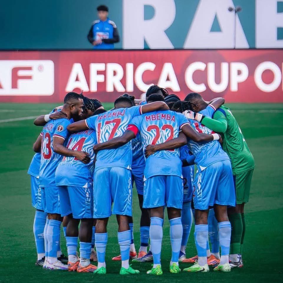 Soccer players in blue jerseys form a tight huddle on the green pitch during the Africa Cup match, arms around each other.