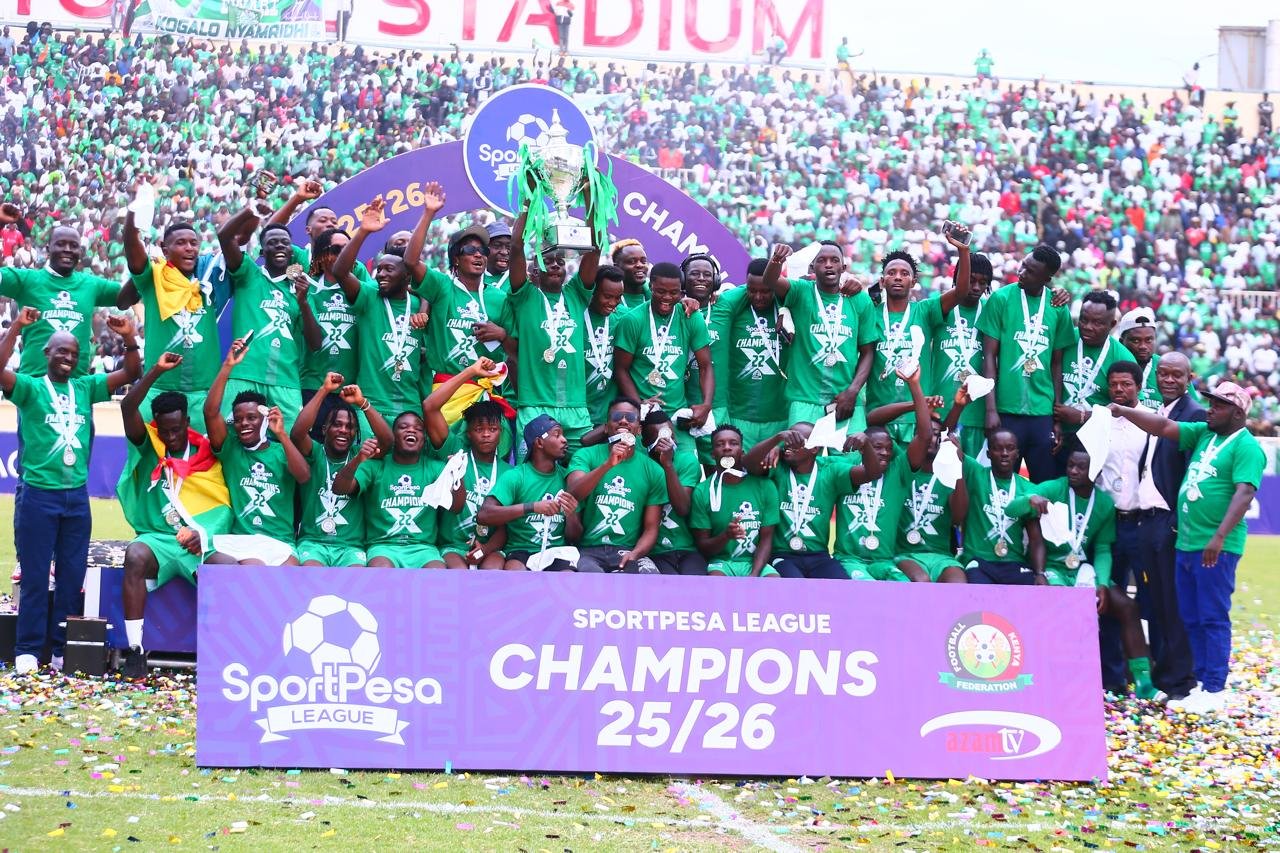 Group of soccer players in green shirts raise a trophy on a stadium field, celebrating as 25/26 SportPesa League champions, with confetti on the ground.