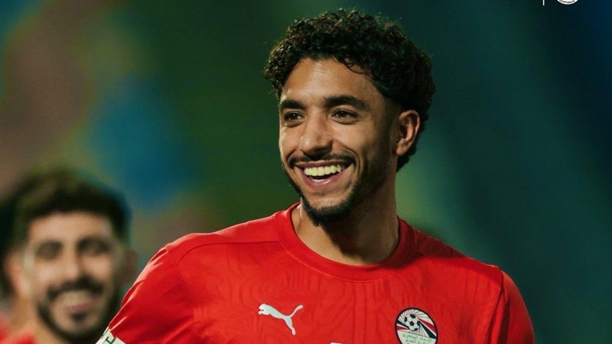 Smiling male soccer player in a red jersey on the field with a blurred crowd behind him.
