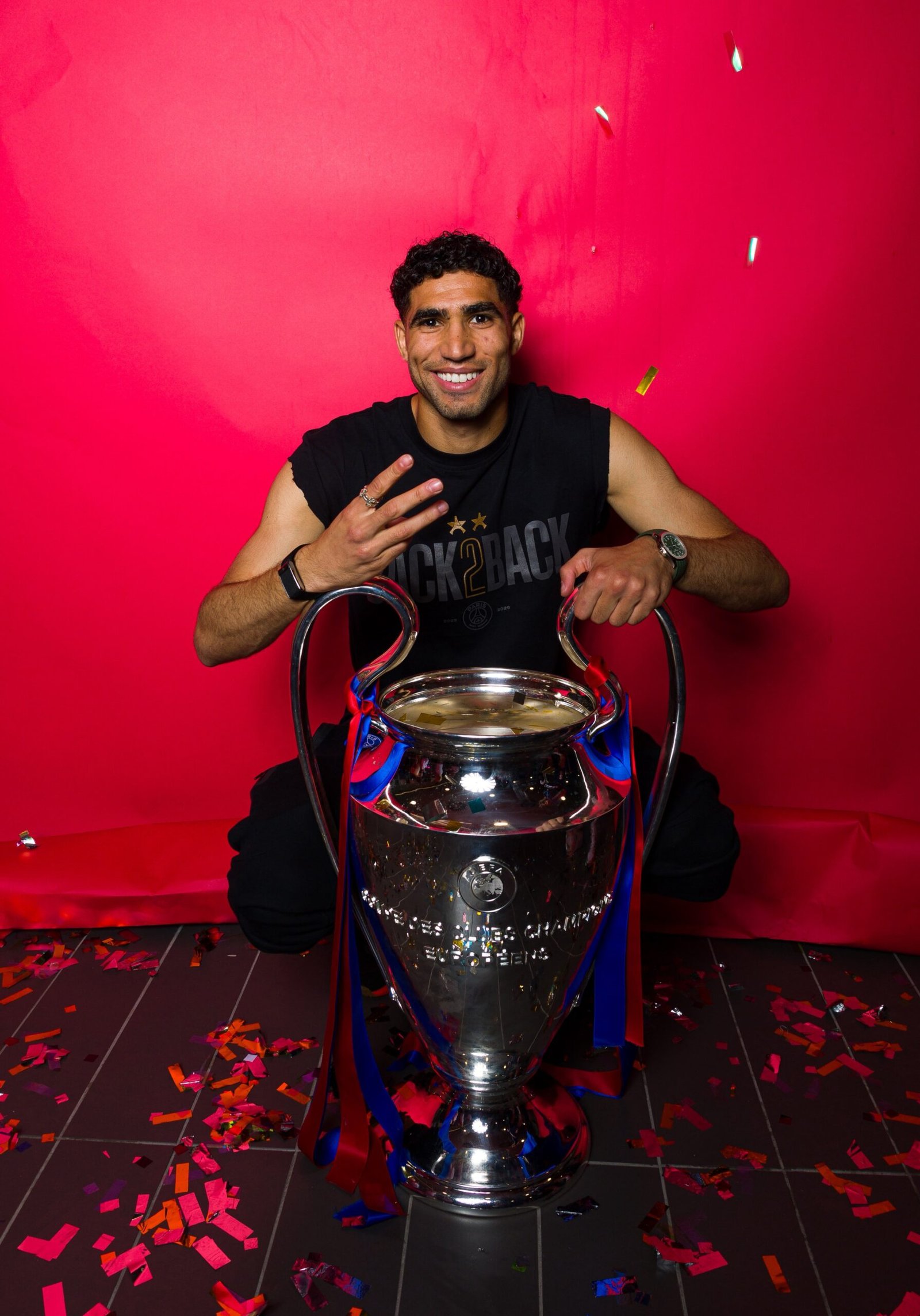 Smiling man in a black sleeveless shirt sits with a large trophy, red backdrop and confetti around.