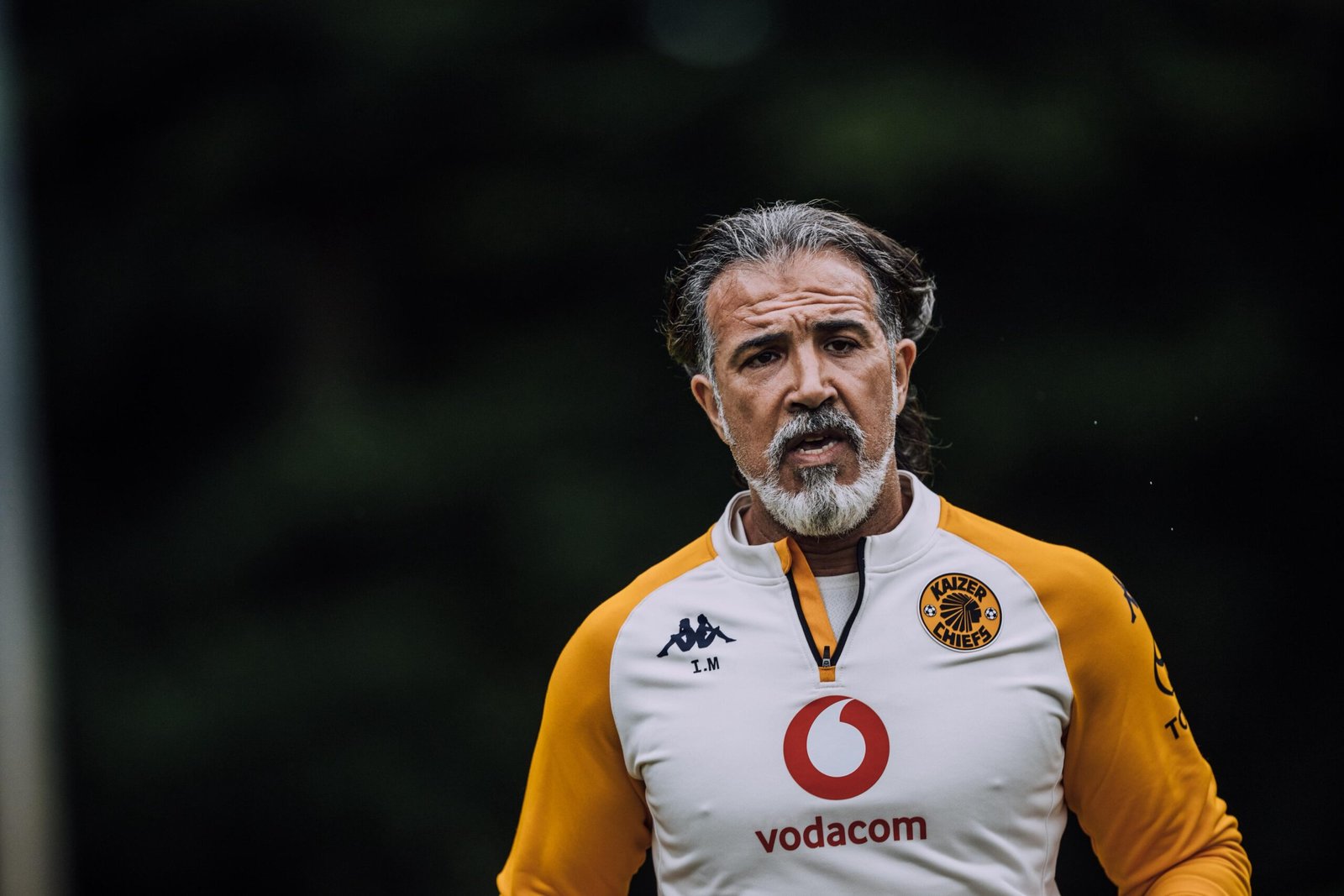 Portrait of a man with gray hair and a white-and-gray beard, wearing an orange and white soccer jersey, facing forward on the field.