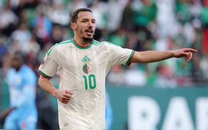 Algerian national team soccer player in white kit with green trim and number 10, gesturing on the field.