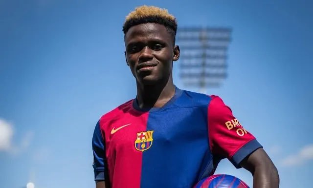 Portrait of a young footballer in a blue and maroon FC Barcelona kit holding a soccer ball, stadium lights in the background on a sunny day.