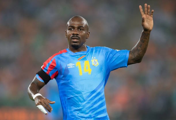 Footballer in a blue kit with number 14 raises his right hand on the field during a match while looking ahead.