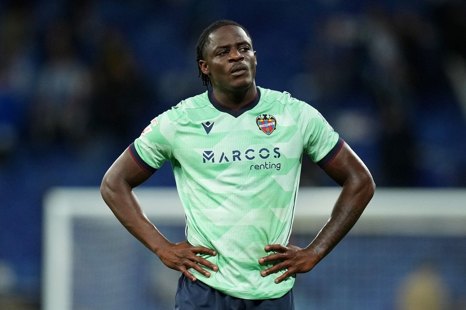 Soccer player in a pale green jersey with crest, standing with hands on hips on the field.