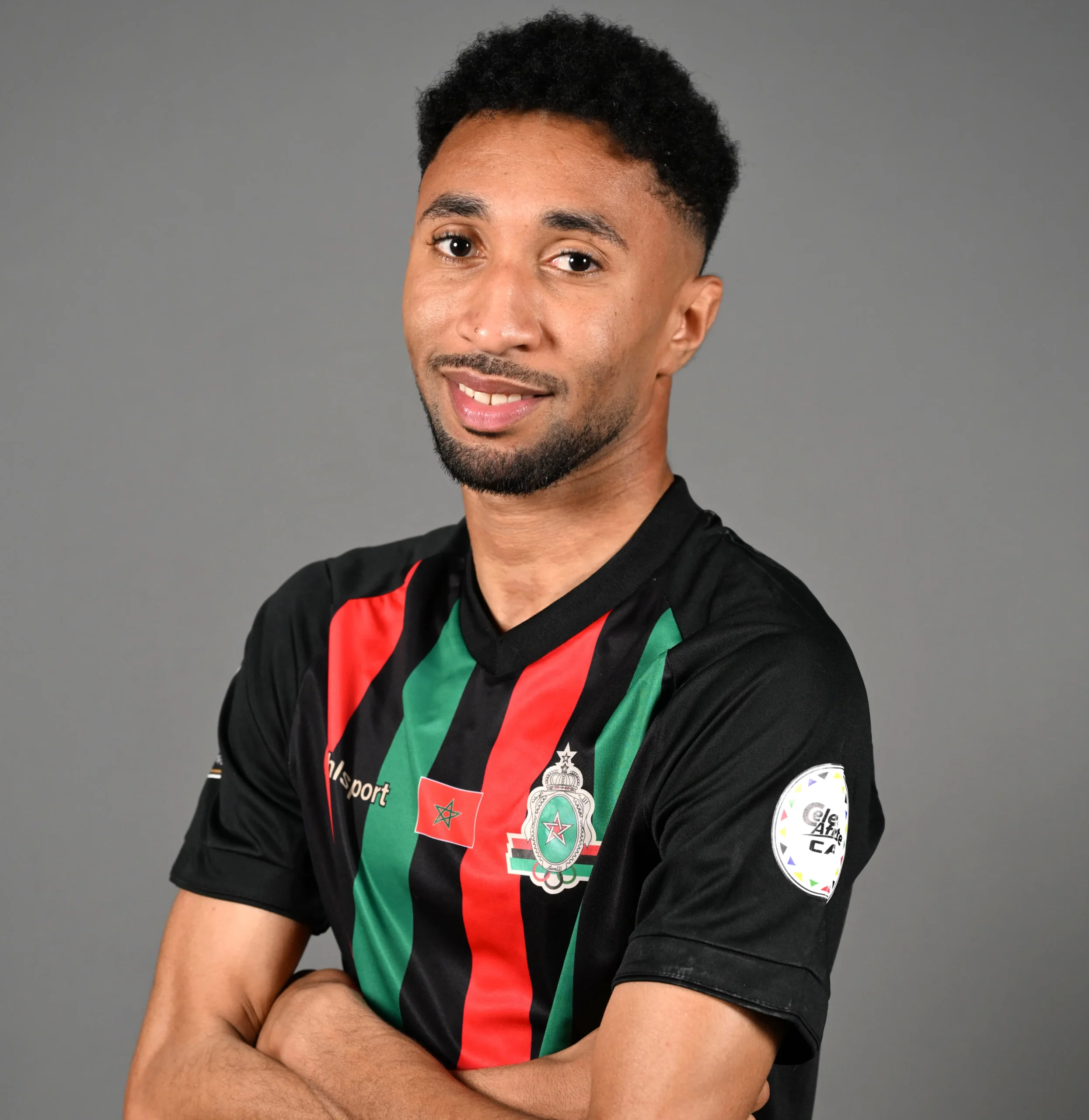 Portrait of a smiling man with short curly hair and a beard, wearing a black sports jersey with red and green diagonal stripes against a gray backdrop.