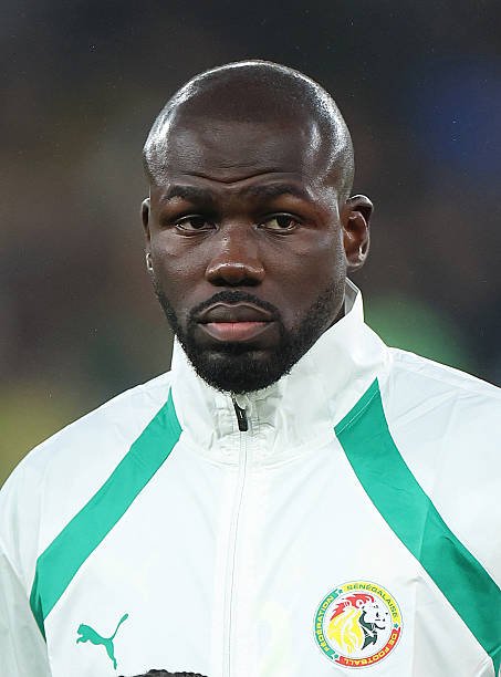 Close-up of a dark-skinned man with a beard wearing a white Senegal training jacket with green stripes, looking focused. Close-up of a dark-skinned man with a beard wearing a white Senegal training jacket with green stripes, looking focused.