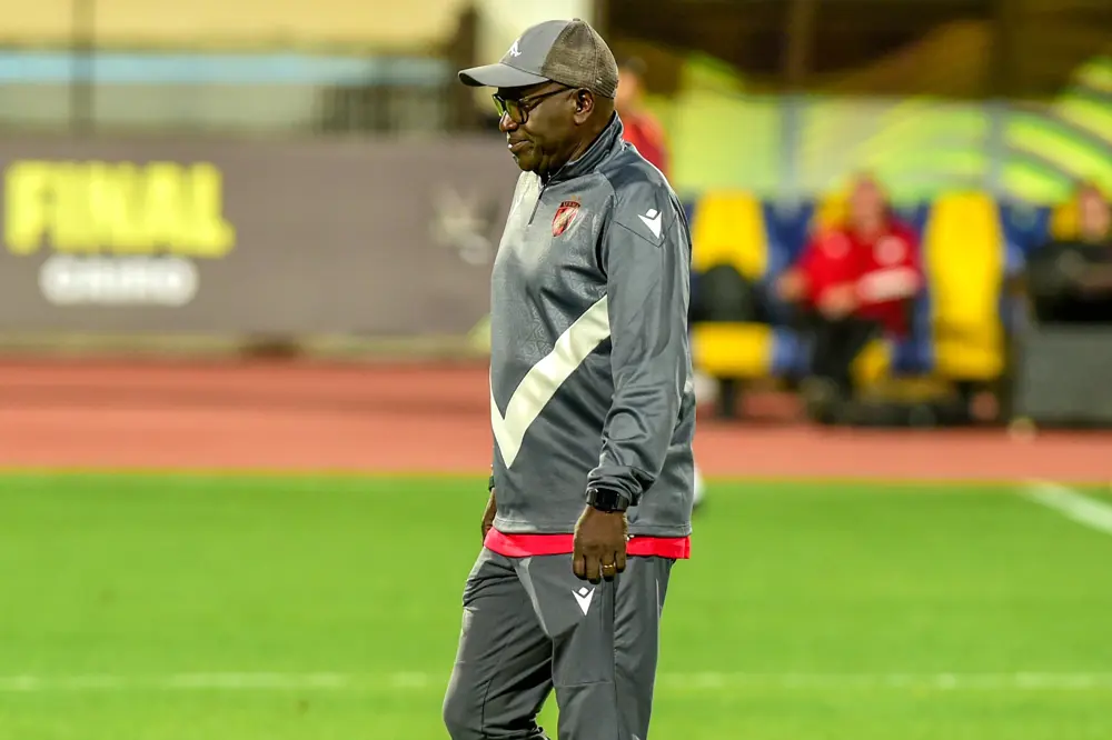 Coach in a gray track suit with a white chevron walks across a football pitch, wearing a cap and glasses, with a blurred bench area and a 'FINAL' sign in the background. Coach in a gray track suit with a white chevron walks across a football pitch, wearing a cap and glasses, with a blurred bench area and a 'FINAL' sign in the background.