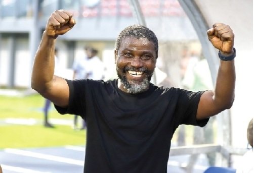Smiling man in a black shirt raises both arms in celebration on a sports field.