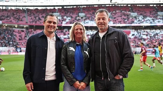 Three adults posing on a soccer field inside a stadium, smiling at the camera.