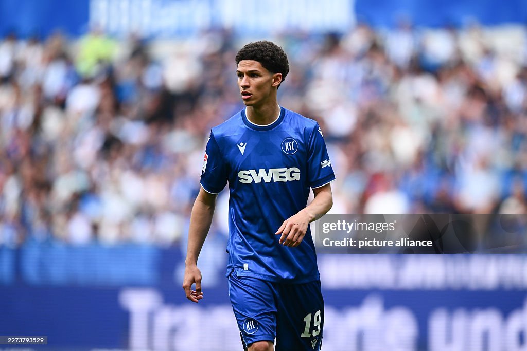 Soccer player in a blue kit with SWEG sponsor walking on the field during a match, stands and crowd behind him.