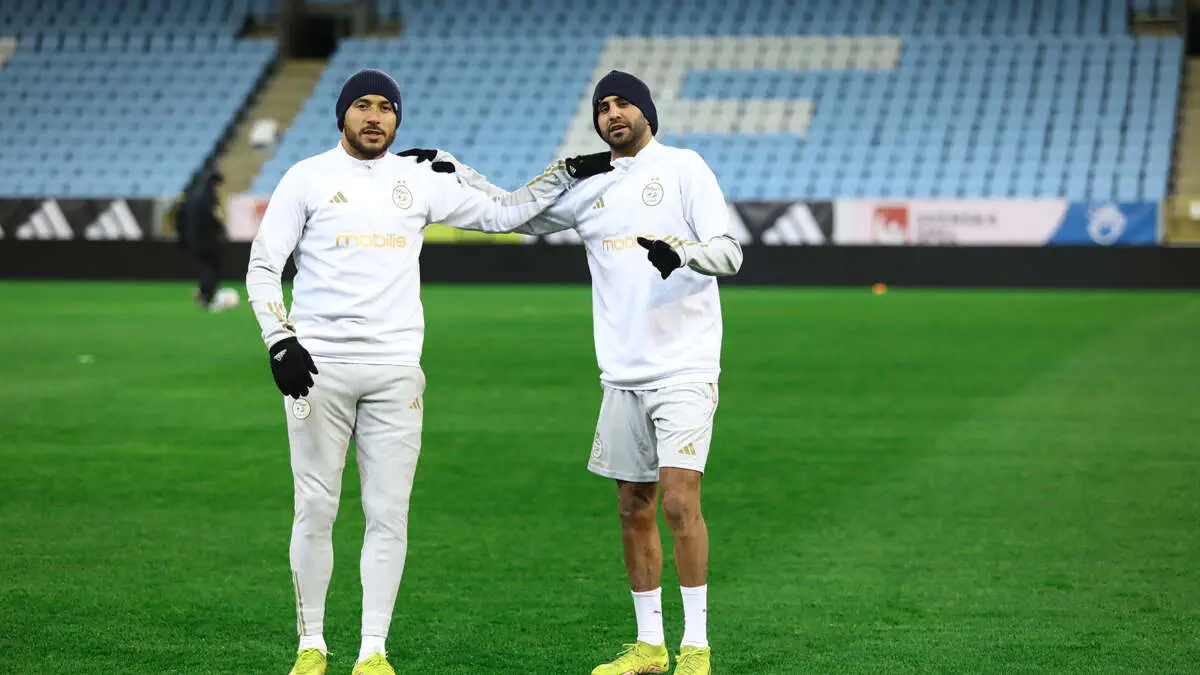 Two soccer players in white training gear pose with arms around each other on a green stadium field. Two soccer players in white training gear pose with arms around each other on a green stadium field.