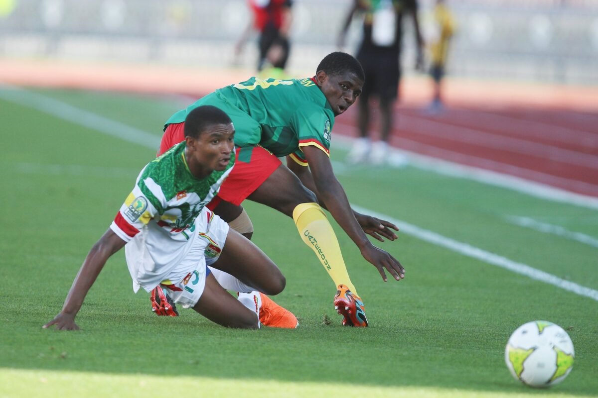 Two soccer players compete for the ball on a green pitch; one in green jersey and yellow socks reaches forward as the ball rolls nearby. Two soccer players compete for the ball on a green pitch; one in green jersey and yellow socks reaches forward as the ball rolls nearby.