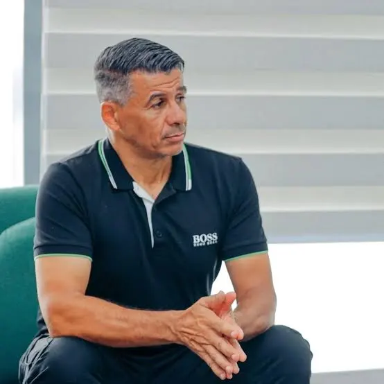 Man in a black polo shirt sits with hands clasped, looking to the left.