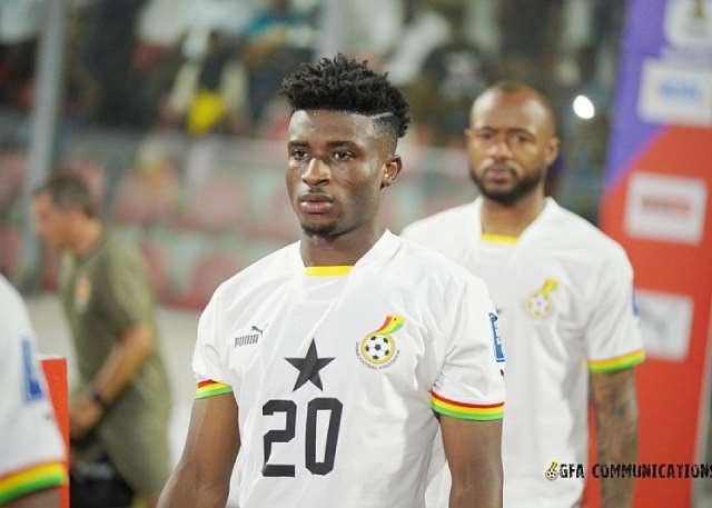 Footballer wearing a white Ghana jersey with number 20 walking on the sidelines, another teammate in the background.