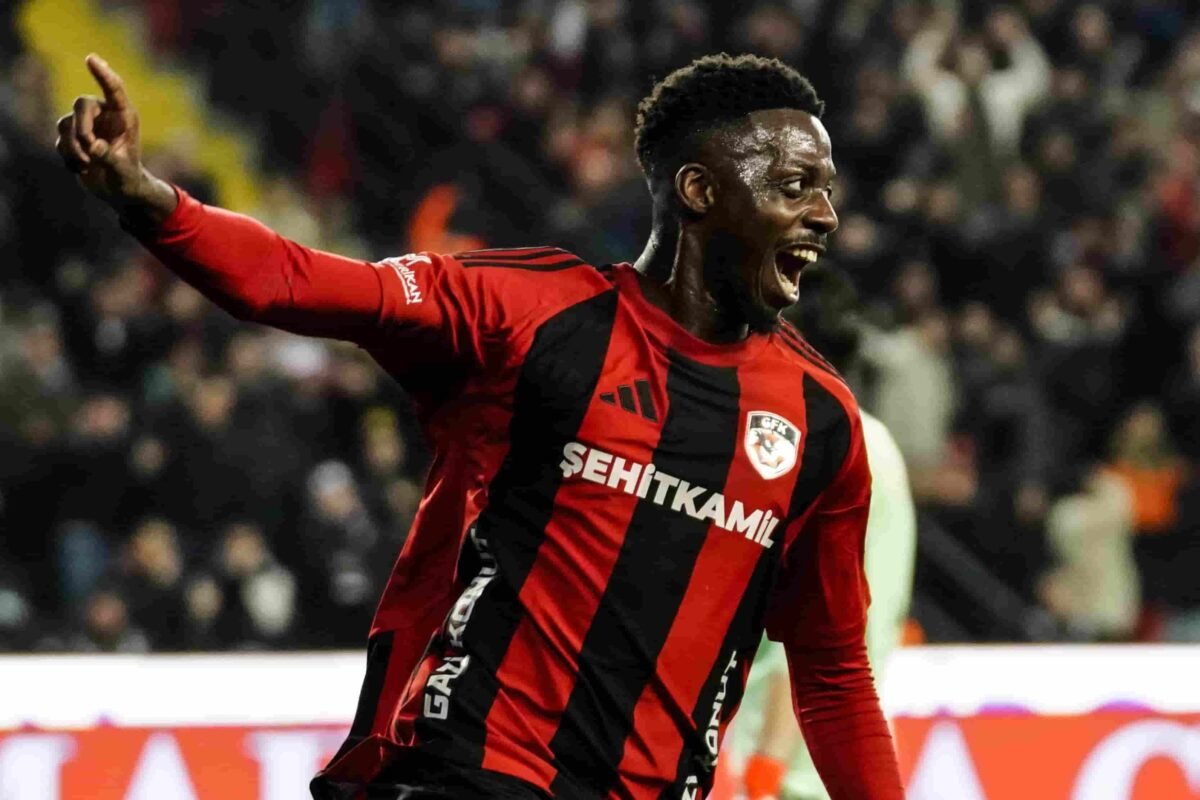 Soccer player in a red-and-black striped jersey celebrates with one arm raised as fans cheer in the stadium.