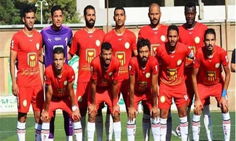 Soccer team in red jerseys posing for a group photo on a green field; goalkeeper in purple stands among them. Soccer team in red jerseys posing for a group photo on a green field; goalkeeper in purple stands among them.