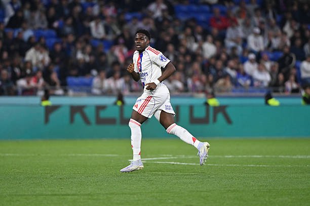 Soccer player in a white kit sprinting on a green pitch during a match, with spectators in the stands behind him.