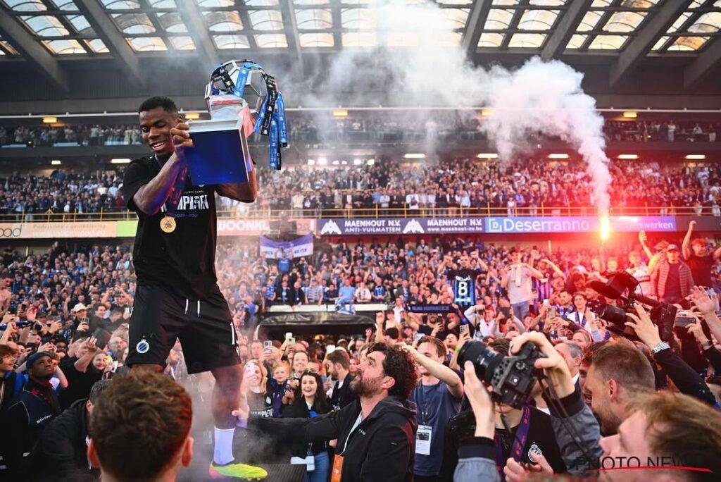 Footballer on a podium lifts a trophy as a cheering stadium crowd watches, with a flare in the air behind him.