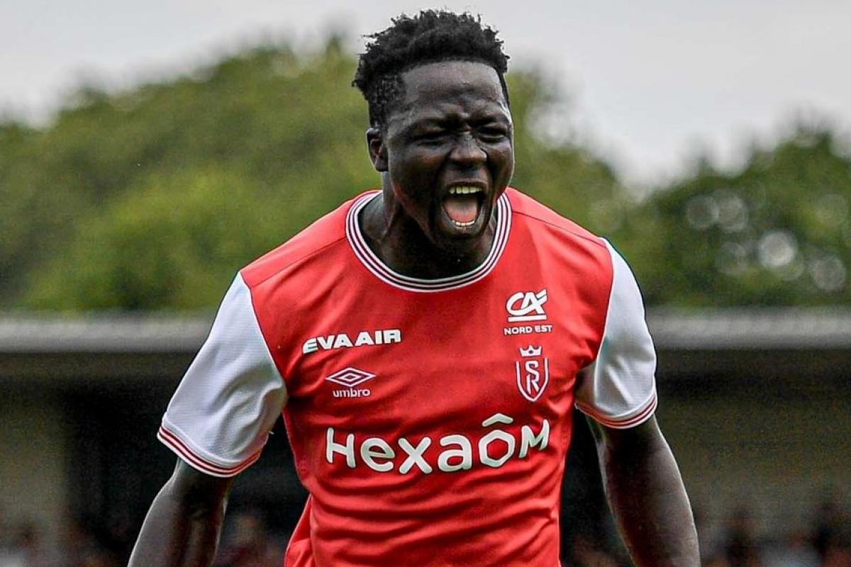 Footballer in a red jersey shouts passionately on the pitch, arms slightly raised.