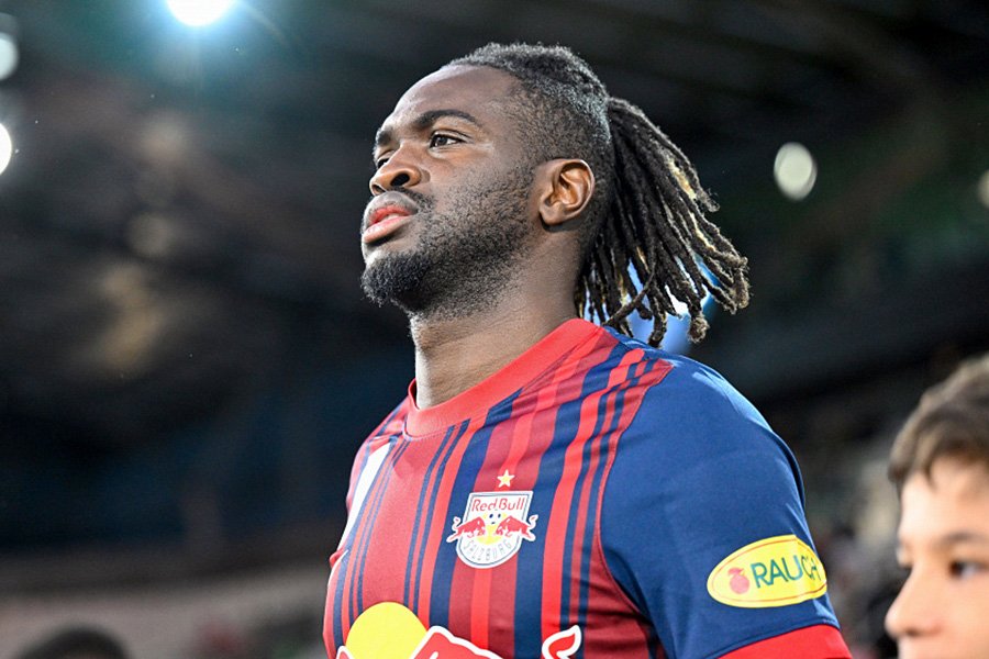 Soccer player with braided hair wearing a Red Bull jersey on the field, looking upward under stadium lights in the background at dusk or nightfall.
