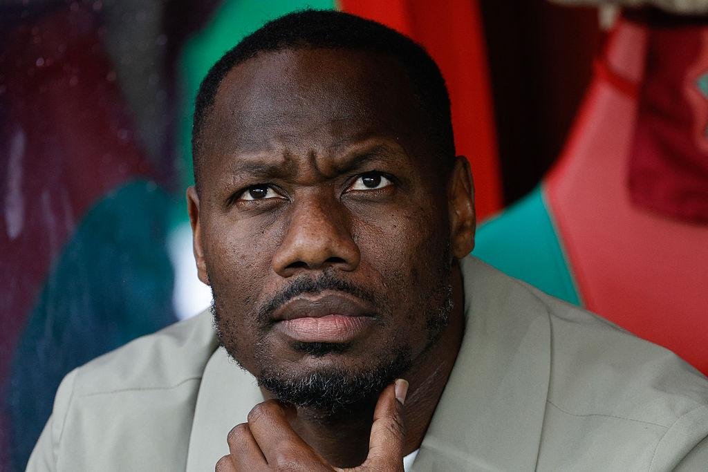 Close-up portrait of a man with a serious, contemplative expression, hand resting on his chin in a colorful background, beige shirt.