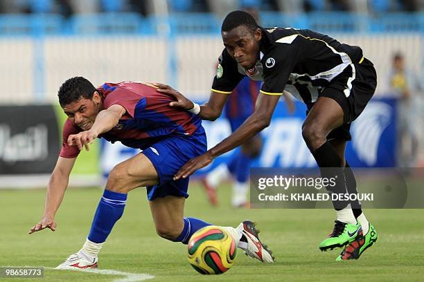 Two football players sprint toward the ball on a green pitch, one in maroon-blue and the other in black kit, mid-challenge for possession.