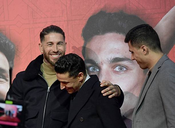 Three men pose together, smiling in front of a red backdrop showing a large portrait; left in a black jacket, middle leaning with arm around another, right in a gray suit.