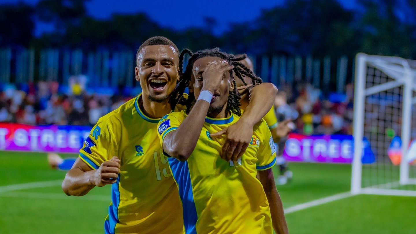 Two soccer players in yellow jerseys celebrate, arms around each other on the field with a cheering crowd behind them.