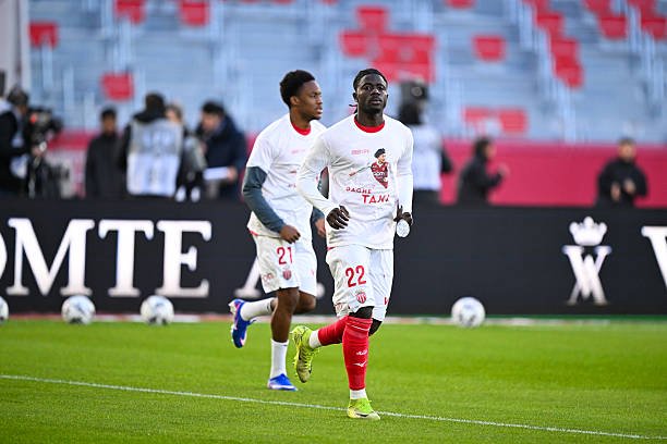 Two soccer players in white and red kits jog on a green pitch during warm-up, with empty stadium seats behind them.