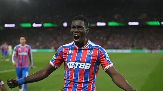 Soccer player in a red-and-blue striped kit celebrates with arms outstretched on a green field.